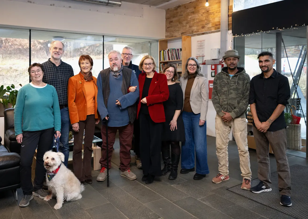 Eine Gruppe von Menschen, einschließlich Männer und Frauen, posiert für ein Foto in Innenräumen. Sie tragen alle Winterkleidung. Ein Hund sitzt auf dem Boden. Hinter ihnen ist eine Glaswand mit Blick nach draußen.