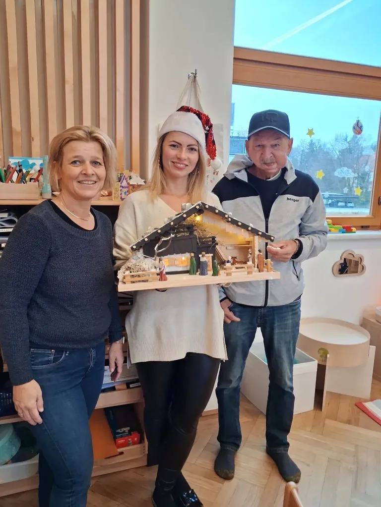 Drei Personen posieren für ein Foto in einem Raum. Die Frau in der Mitte hält eine kleine hölzerne Krippe. Sie alle lächeln für die Kamera. Hinter ihnen steht ein Regal und ein Fenster mit Blick auf Bäume.
