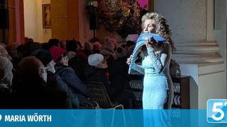 Eine Frau in einem blauen Kleid spielt vor einem sitzenden Publikum eine Harfe, mit einer Treppe und einer dekorierten Wand im Hintergrund.