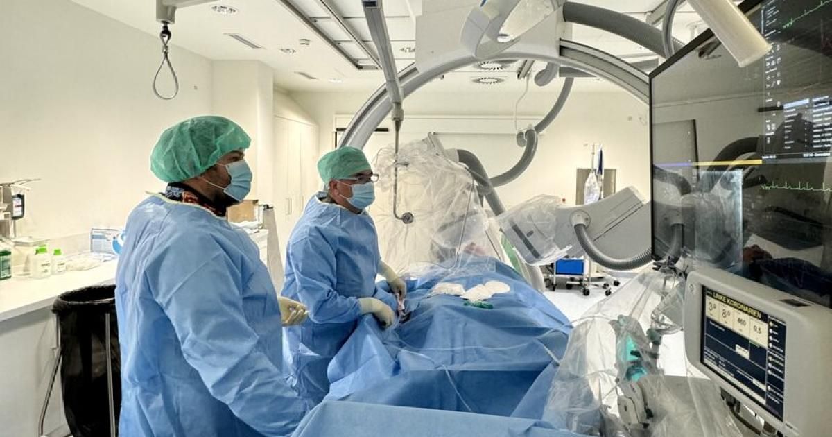 Zwei medizinische Fachkräfte in Operationskleidung, einschließlich Masken und Haarnetzen, operieren einen Patienten in einem Krankenhauszimmer. Sie stehen über einem Operationstisch mit Operationstüchern und moderner medizinischer Ausrüstung.