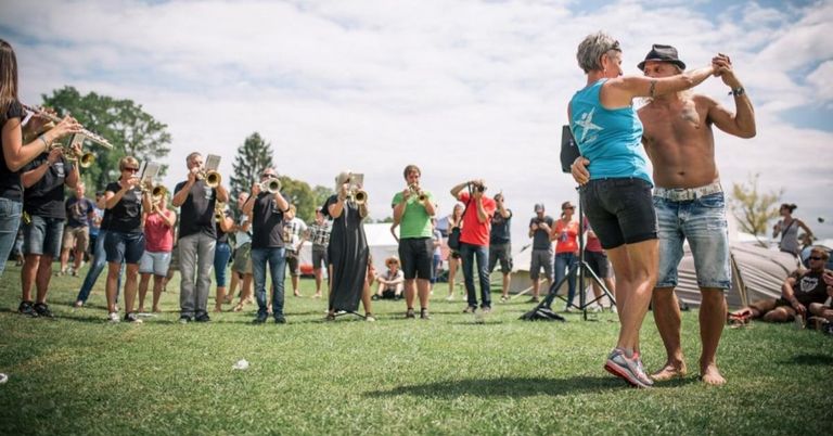 Eine Gruppe von Menschen steht auf einem Rasenfeld, einige spielen Trompeten, während andere zuschauen. Eine Frau umarmt einen Mann vor ihnen.
