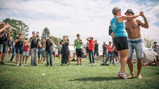 Eine Gruppe von Menschen steht auf einem Rasenfeld, einige spielen Trompeten, während andere zuschauen. Eine Frau umarmt einen Mann vor ihnen.