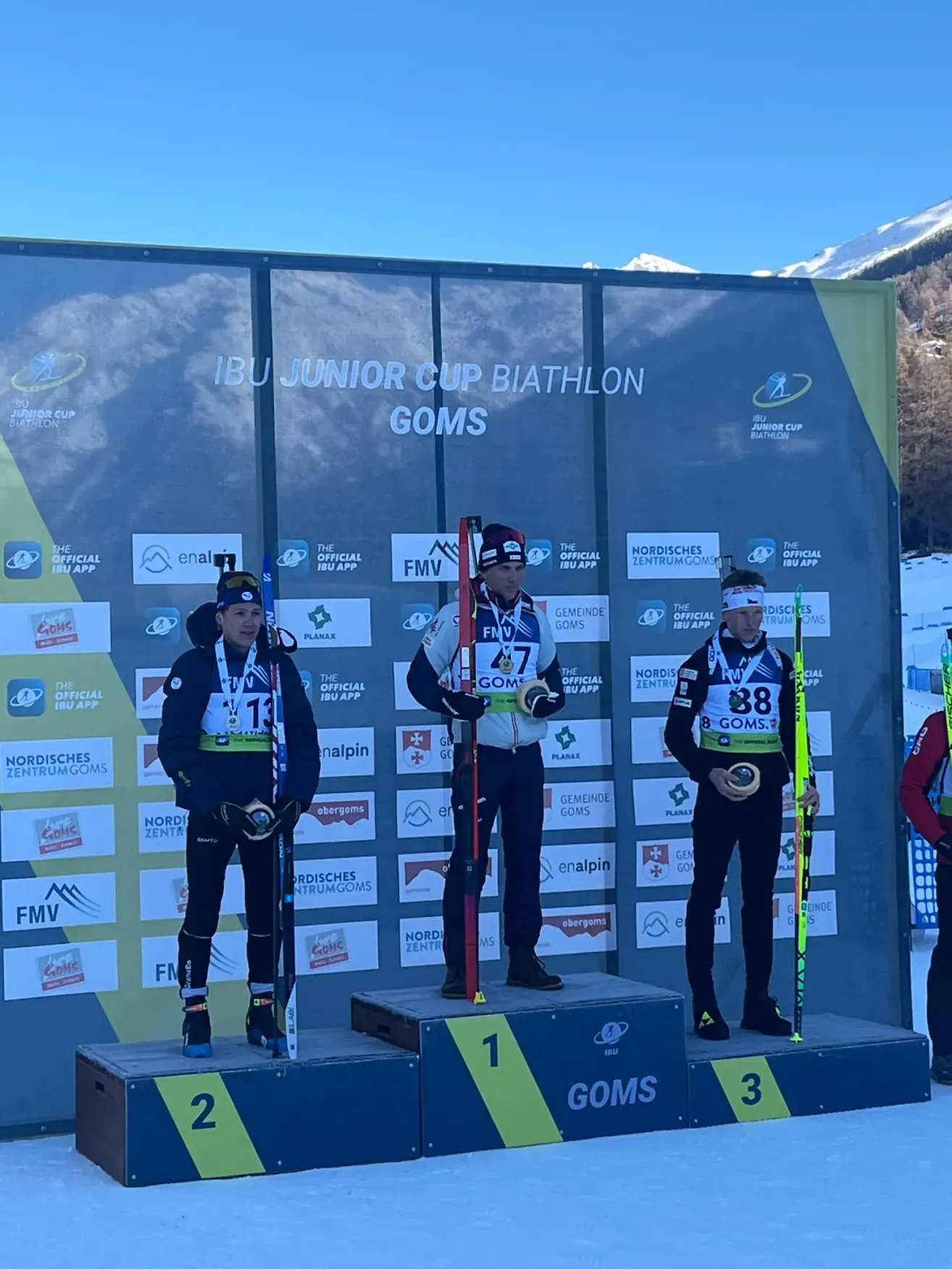 Drei Biathleten stehen auf einem Podium mit einem Hintergrund, auf dem Ibu Junior Cup Biathlon in Goms steht. Jeder Athlet trägt eine Medaille und hält einen Skistock. Der erste Platz hat die Nummer 1 auf ihrem Podium.