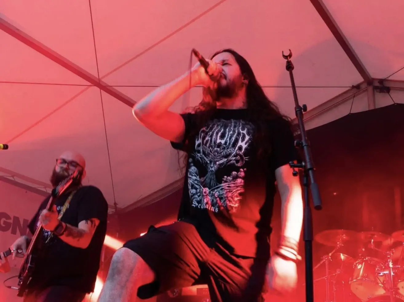 Ein Mann mit langen Haaren und Bart steht auf der Bühne und singt in ein Mikrofon. Ein anderer Mann spielt Gitarre neben ihm. Die Bühne ist mit roten Lichtern beleuchtet.