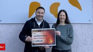 Ein Mann und eine Frau stehen nebeneinander und halten einen Spendenscheck. Sie lächeln beide und wirken glücklich. Der Mann trägt eine schwarze Jacke, und die Frau trägt einen grünen Pullover.