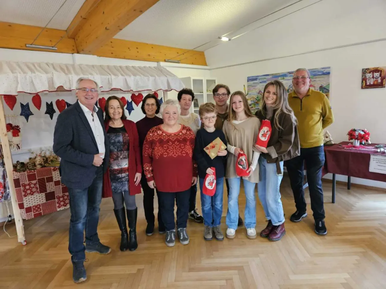 Eine Gruppe von Menschen, einschließlich Erwachsener und Kindern, steht lächelnd und mit Geschenktüten in der Hand in einem Raum. Hinter ihnen befindet sich ein weißer Vorhang mit Herzdesigns.