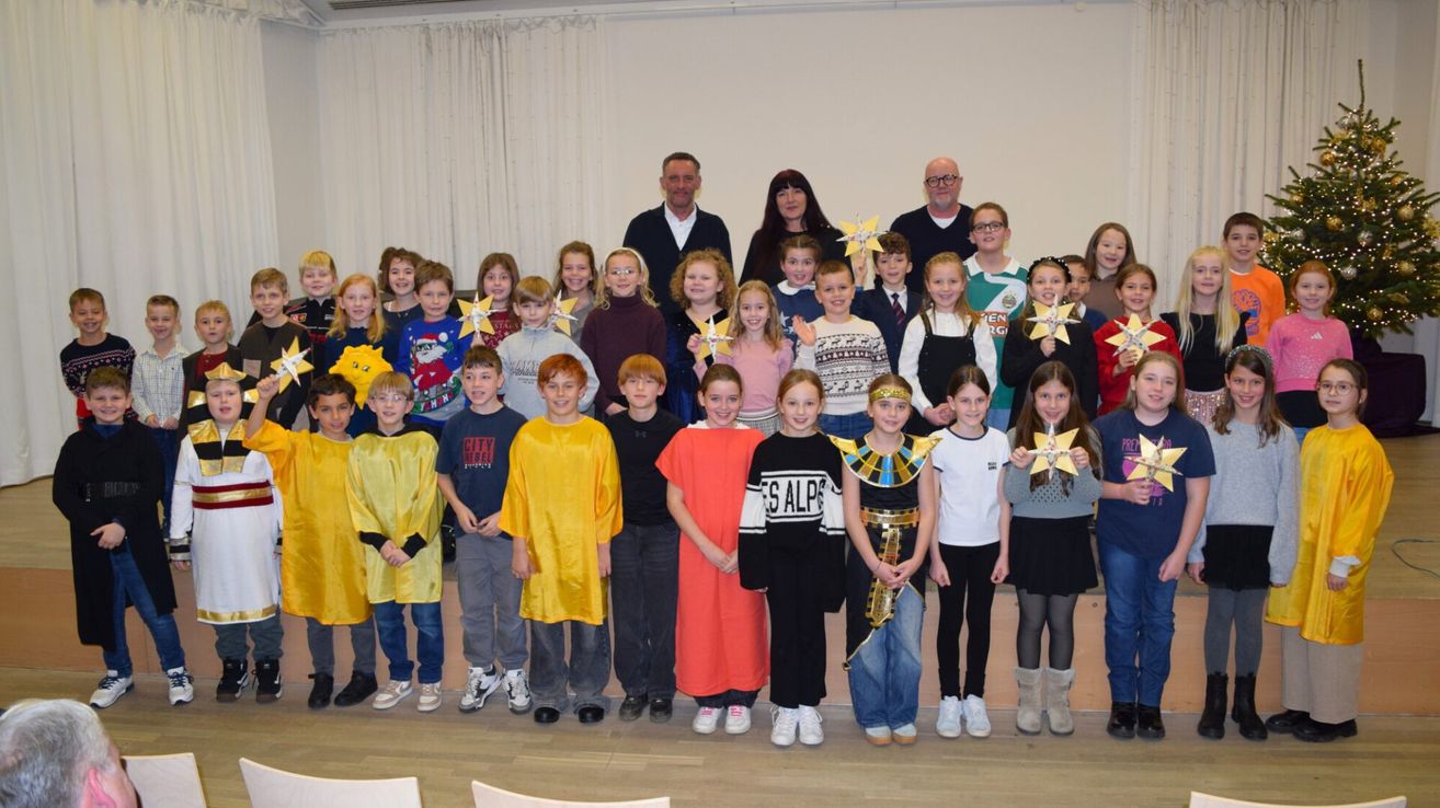 Eine Gruppe von Kindern in verschiedenen Kostümen, einige halten Sterne, posiert für ein Foto mit zwei Erwachsenen in einem Klassenzimmer.