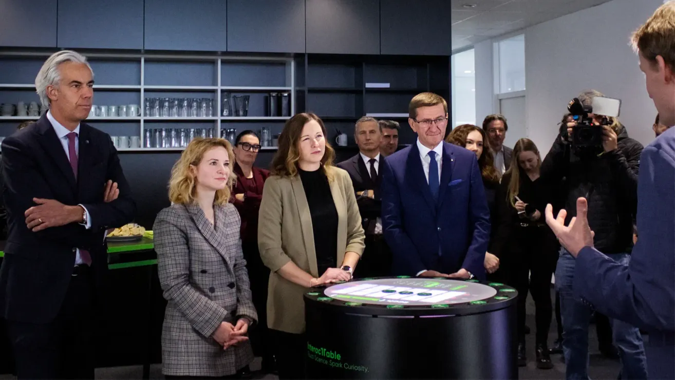 Mehrere Personen stehen um einen Tisch herum, wobei zwei Frauen davor stehen. Der Mann im blauen Anzug lächelt und schaut in die Kamera. Dahinter befindet sich ein Regal mit Gläsern und eine weiße Tür.