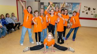 Eine Gruppe junger Mädchen in orangefarbenen T-Shirts posiert für ein Foto in einem Klassenzimmer. Einige stehen, andere sitzen oder liegen. Der Raum ist mit Postern und Dekorationen an den Wänden geschmückt.