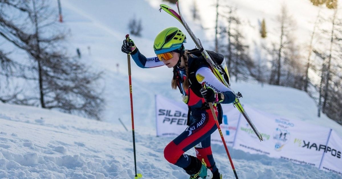 Eine Skifahrerin in einem grünen Helm fährt bergab auf einem verschneiten Hang und trägt Skier auf den Schultern. Sie trägt einen roten und weißen Rennanzug und hält Skistöcke. Hinter ihr sind Banner und Bäume zu sehen.