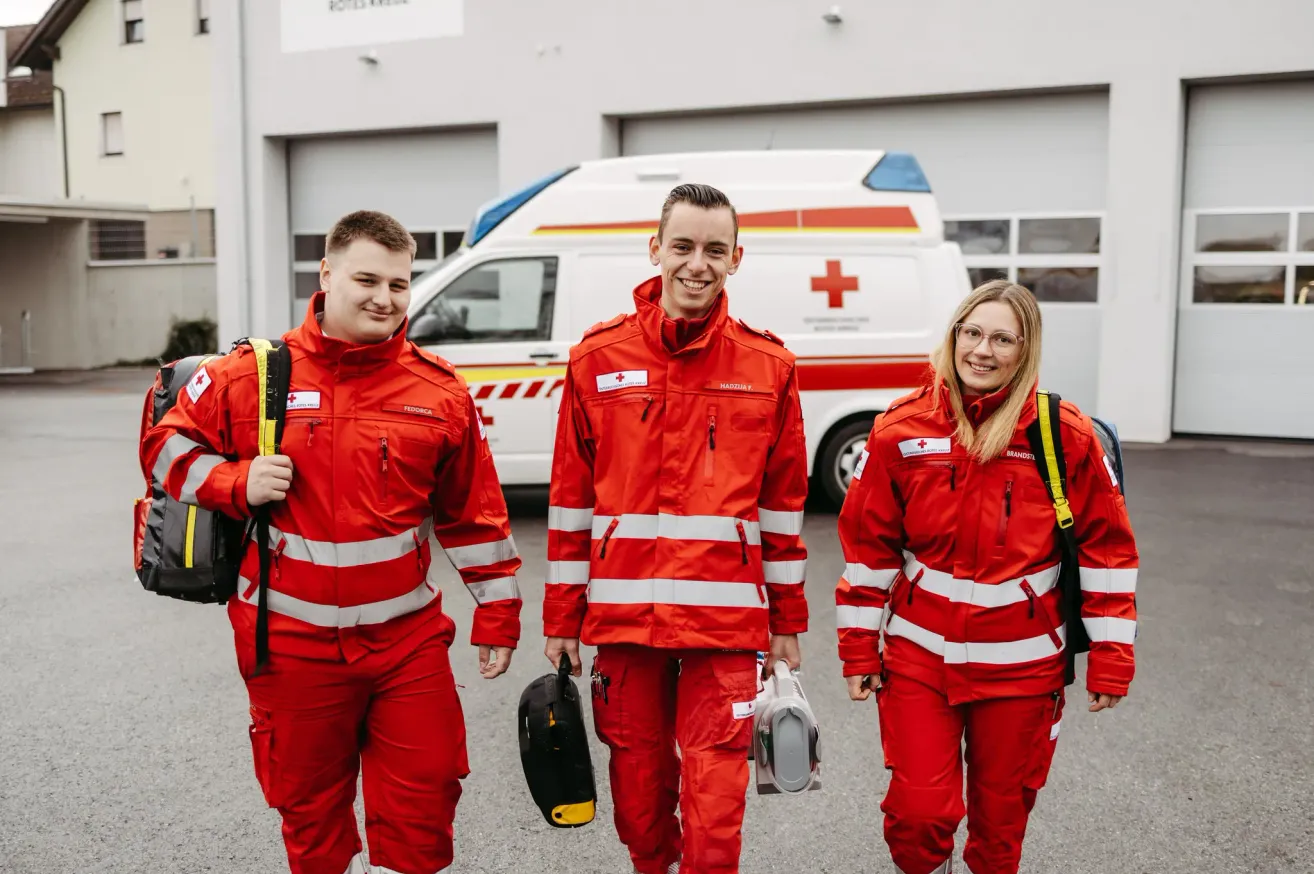 Drei Personen in roten Uniformen mit reflektierenden Streifen gehen nach draußen. Sie lächeln und scheinen für ein Foto zu posieren. Der Mann auf der linken Seite trägt einen Rucksack. Ein weißer Krankenwagen ist hinter ihnen geparkt.