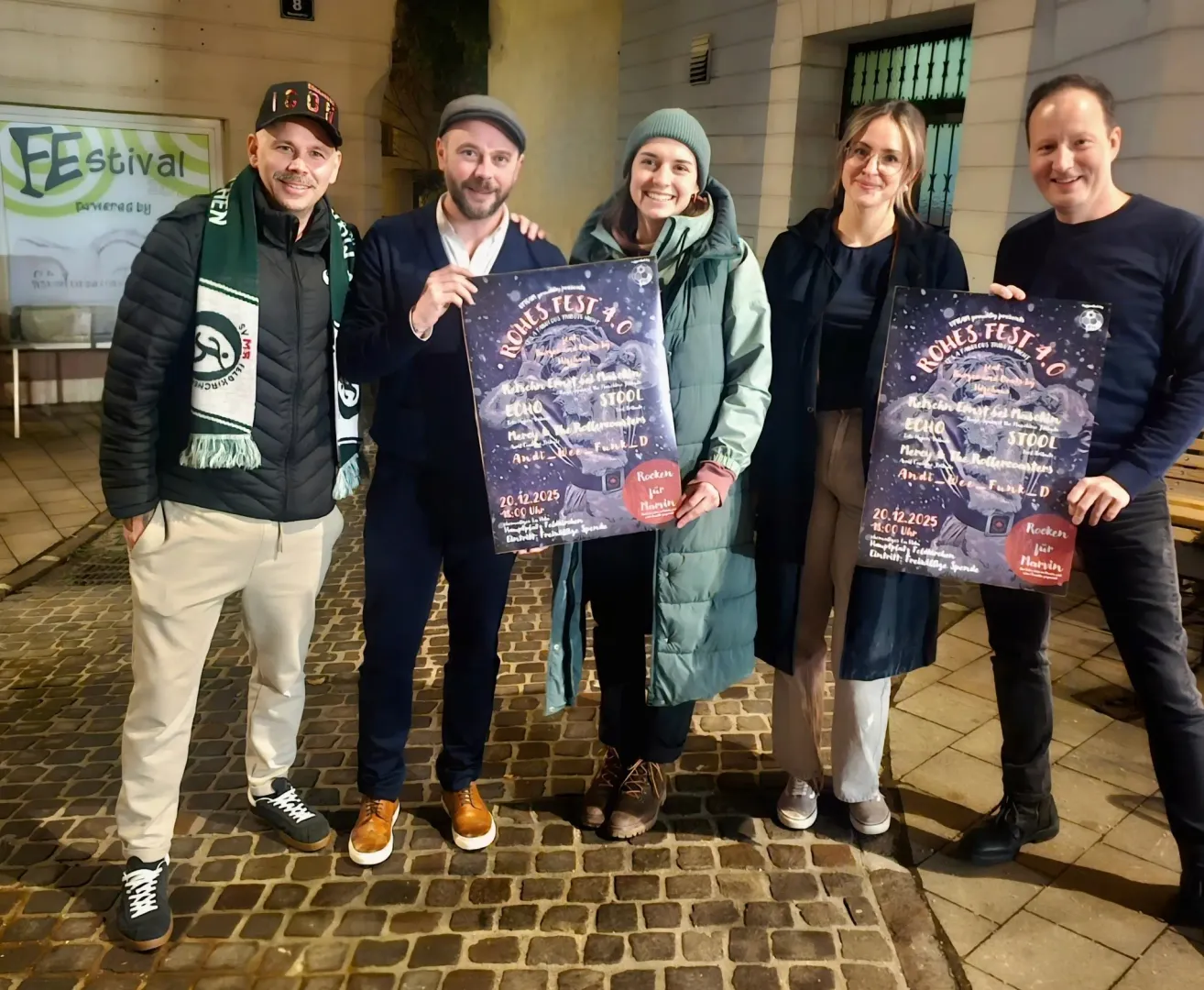Fünf Menschen stehen lächelnd draußen und halten Plakate hoch. Zwei Frauen und drei Männer, einer mit einem Schal, ein anderer mit einem Hut. Ziegelboden und ein Gebäude im Hintergrund.