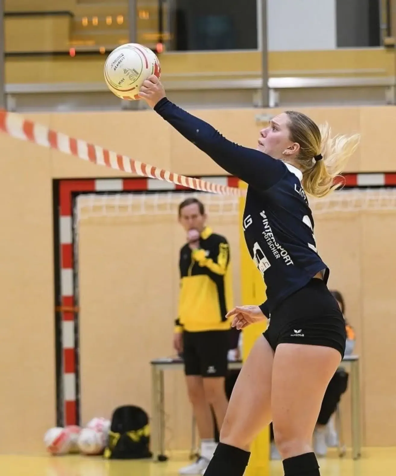 Eine Frau in einem schwarzen und gelben Trikot spielt Volleyball und springt, um den Ball zu schlagen. Im unscharfen Hintergrund sieht man ein Netz und eine Person in einem gelben Trikot.