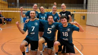 Eine Gruppe Volleyballspieler in passenden Uniformen posiert für ein Foto auf dem Spielfeld, wobei die Nummern 7, 14, 5 und 18 sichtbar sind. Sie befinden sich in einer Sporthalle mit einem Netz im Hintergrund.