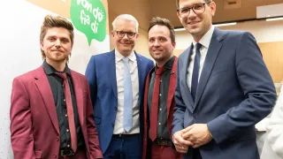 Vier Männer in Anzügen und Krawatten posieren für ein Foto vor einem Banner, auf dem in grünen Sprechblasen 'von do' steht.