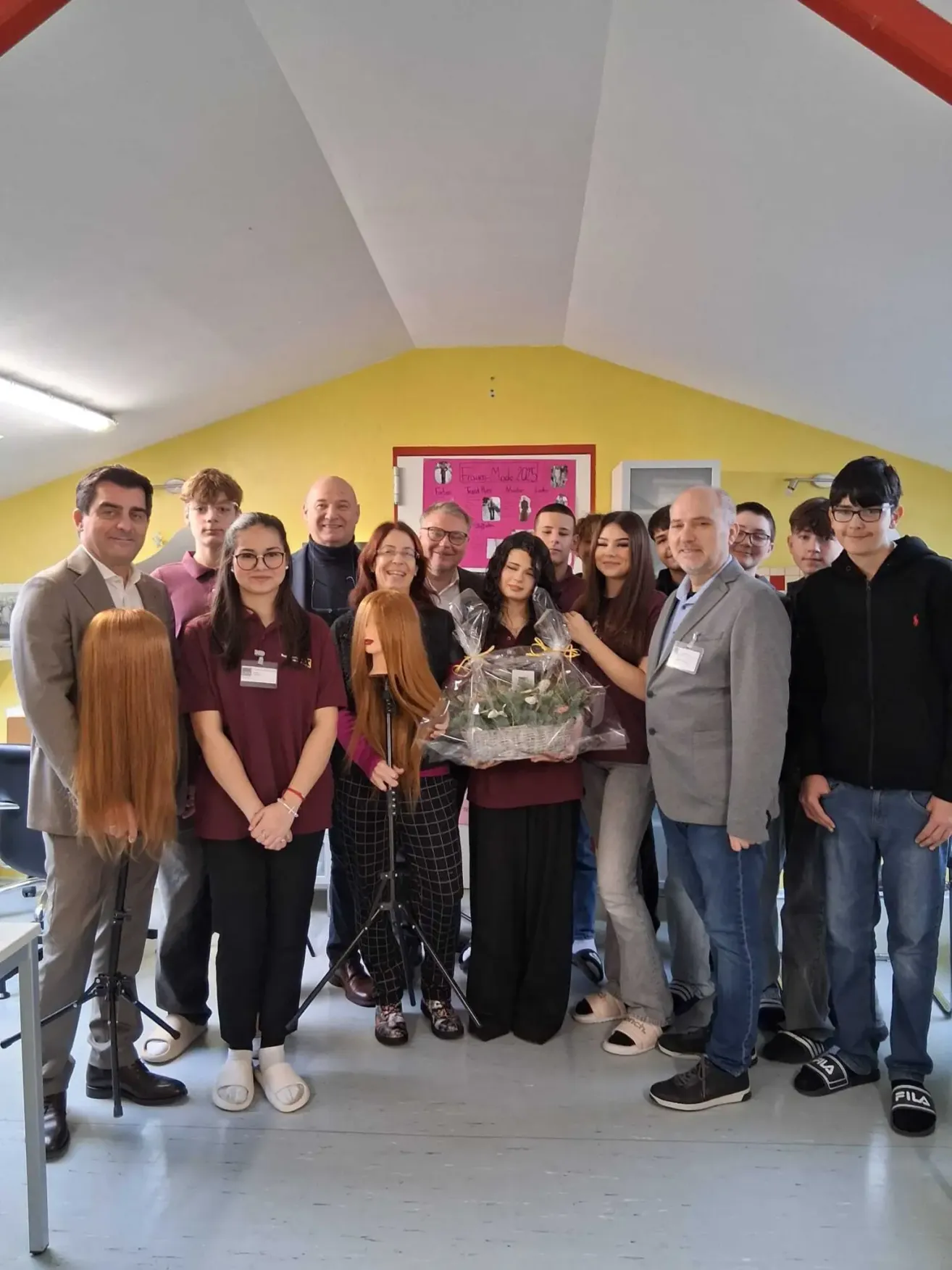 Eine Gruppe von Personen steht in einem Raum zusammen, einige in formeller Kleidung. Eine Frau in der Mitte hält einen Blumenstrauß. Im Hintergrund sind Puppen und Stühle zu sehen, und ein rosa Poster ist an der Wand zu erkennen.