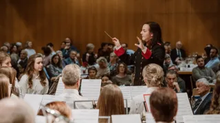 Eine Frau in einer schwarz-roten Uniform dirigiert ein Orchester, mit einer Menge von Menschen im Hintergrund, einige halten Notenblätter.