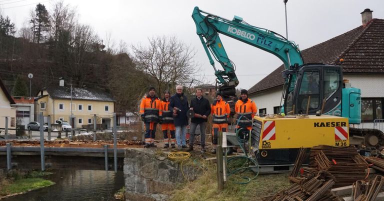Eine Gruppe von Arbeitern in Warnwesten steht neben einer Brücke mit einem großen Bagger im Hintergrund.