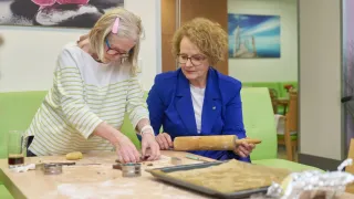 Zwei ältere Frauen arbeiten in einem Raum zusammen, eine hält einen Nudelholz, während die andere mit Teig arbeitet. Es gibt einen Tisch mit Backutensilien und eine Tablett mit Teig. Es gibt Stühle und ein Gemälde an der Wand.