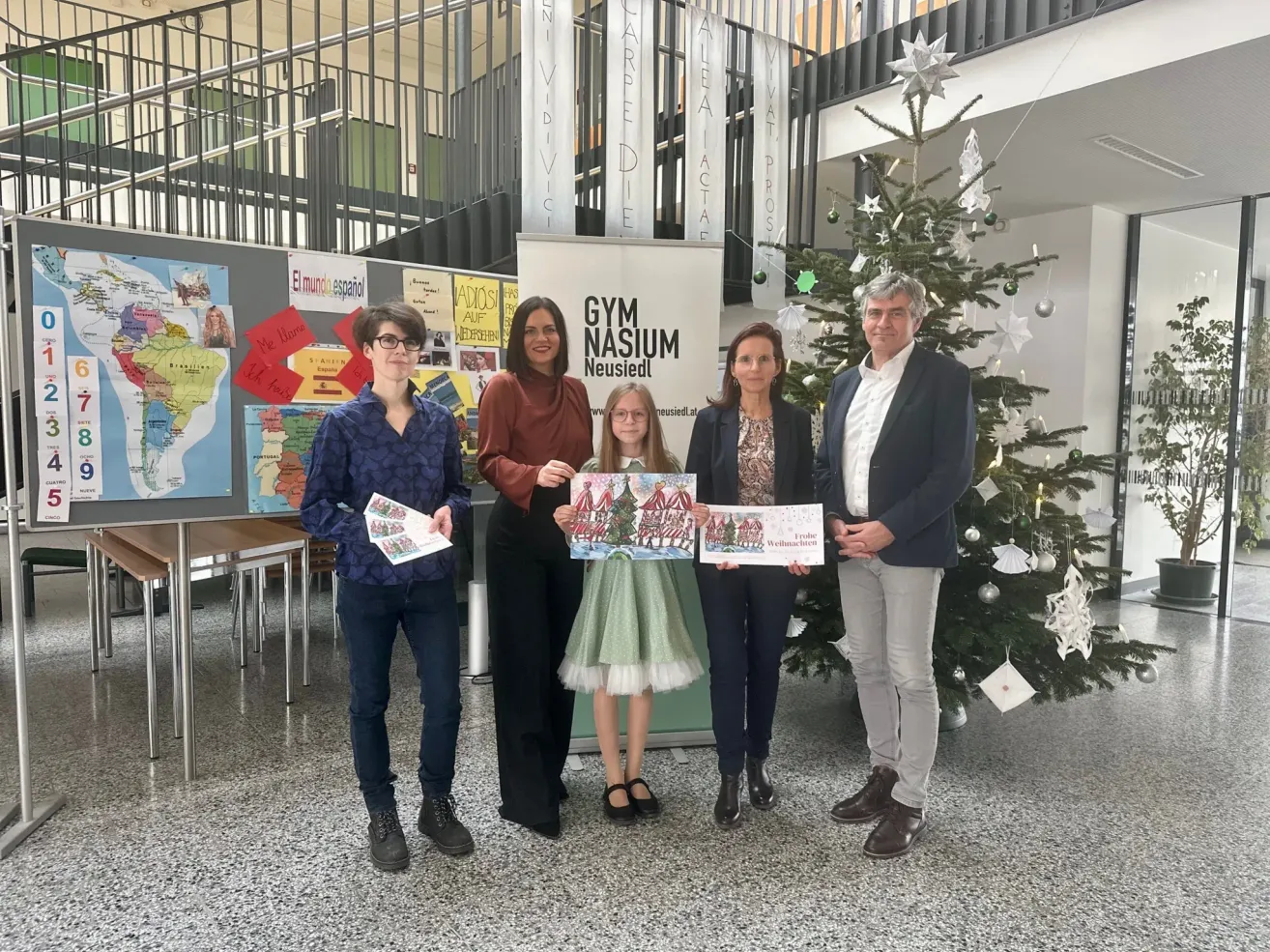 Fünf Personen stehen vor einem Weihnachtsbaum. Sie halten Weihnachtskarten in den Händen. Hinter ihnen befindet sich eine Karte an der Wand.