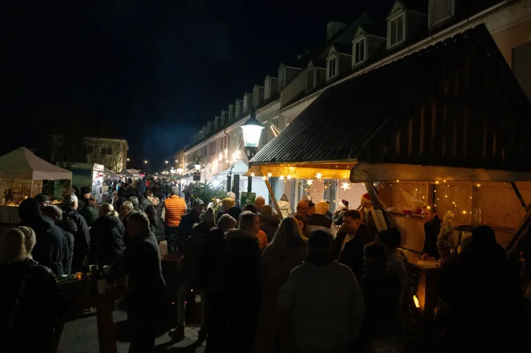 Ein geschäftiger Nachtmarkt in der Dämmerung mit vielen Menschen, die Stände erkunden, einige mit Mützen und Winterkleidung. Eine Straßenlaterne und ein festlich dekorierter Zelt mit leuchtenden Lichtern sind sichtbar.