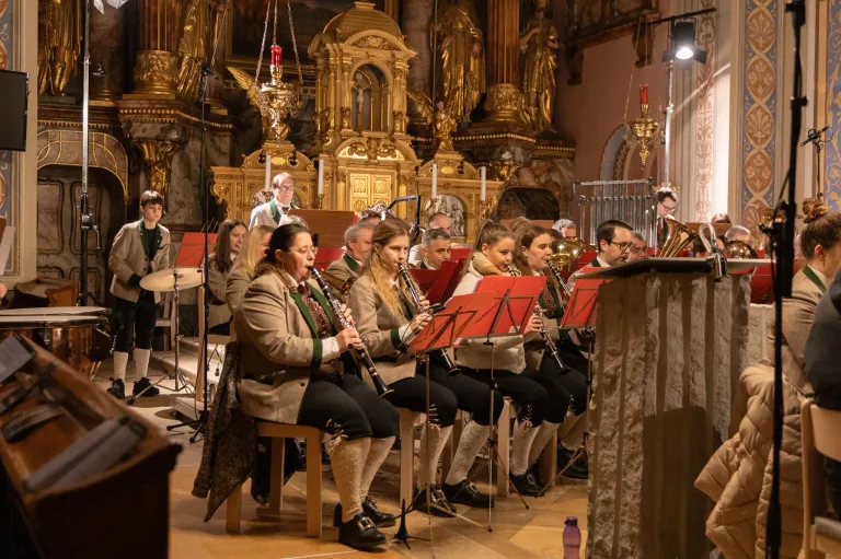 Eine Gruppe von Musikern spielt in einer Kirche, Klarinetten und Trompeten. Ein Mann steht am Kanzel. Mehrere Leute sitzen auf Stühlen, andere stehen hinter ihnen. Der Bereich ist mit goldenen Statuen geschmückt.