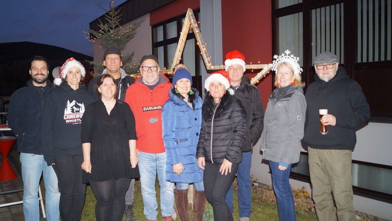 Eine Gruppe Erwachsener, gekleidet in Winterkleidung, steht im Freien vor einem roten Gebäude und lächelt für ein Foto. Sie stehen neben einem Weihnachtsbaum und einer sternförmigen Dekoration.
