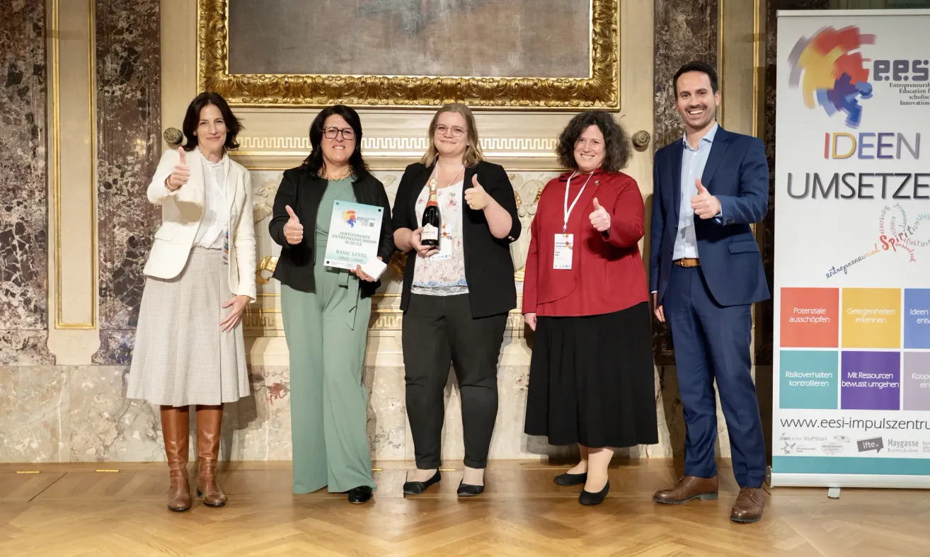 Fünf Personen stehen auf einer Bühne, lächeln und posieren für ein Foto. Eine Frau hält ein Zertifikat, eine andere eine Flasche Champagner. Sie tragen formelle Kleidung, ein Mann einen Anzug. Hinter ihnen befindet sich eine Wand mit einem großen Gemälde.