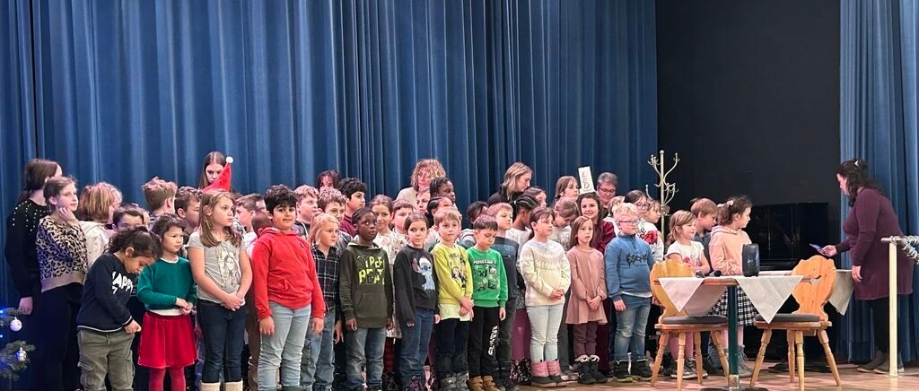 Eine Gruppe von Kindern steht auf einer Bühne vor einem blauen Vorhang, möglicherweise bei einer Schulveranstaltung. Einige Kinder tragen Winterkleidung und eine Brille. Ein paar Erwachsene stehen im Hintergrund.