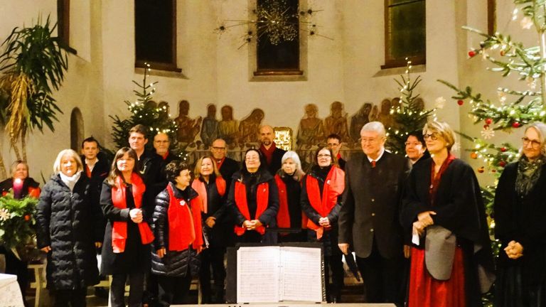 Ein Chor steht in einer Kirche mit roten Gewändern, in der Nähe eines Mannes in einem schwarzen Anzug, mit Weihnachtsbäumen und einem Gemälde im Hintergrund.