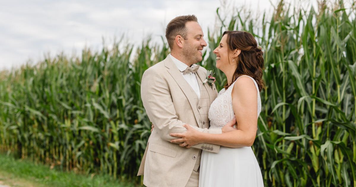 Ein Mann und eine Frau stehen lächelnd vor einem hohen Maisfeld an einem bewölkten Tag.