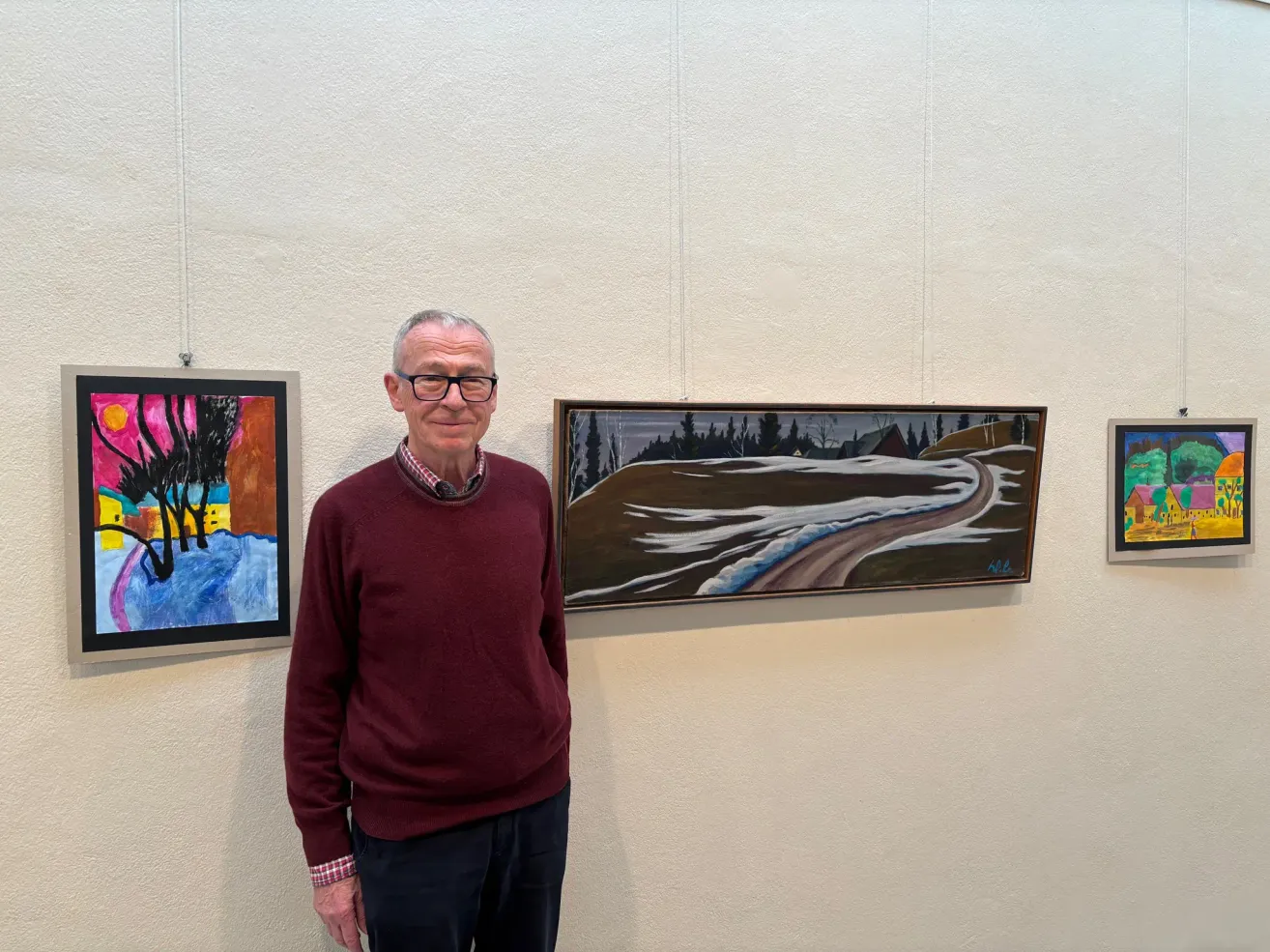 Ein älterer Mann in einem roten Pullover steht vor einer weißen Wand mit zwei gerahmten Gemälden, die daran hängen.