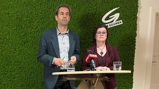 Zwei Personen, ein Mann und eine Frau, stehen hinter einem Podium mit Mikrofonen. Der Mann hält einen grünen Stift, und beide haben Wassergläser auf dem Tisch. Sie stehen vor einer grünen Wand mit dem Wort 'Gruene' darauf.