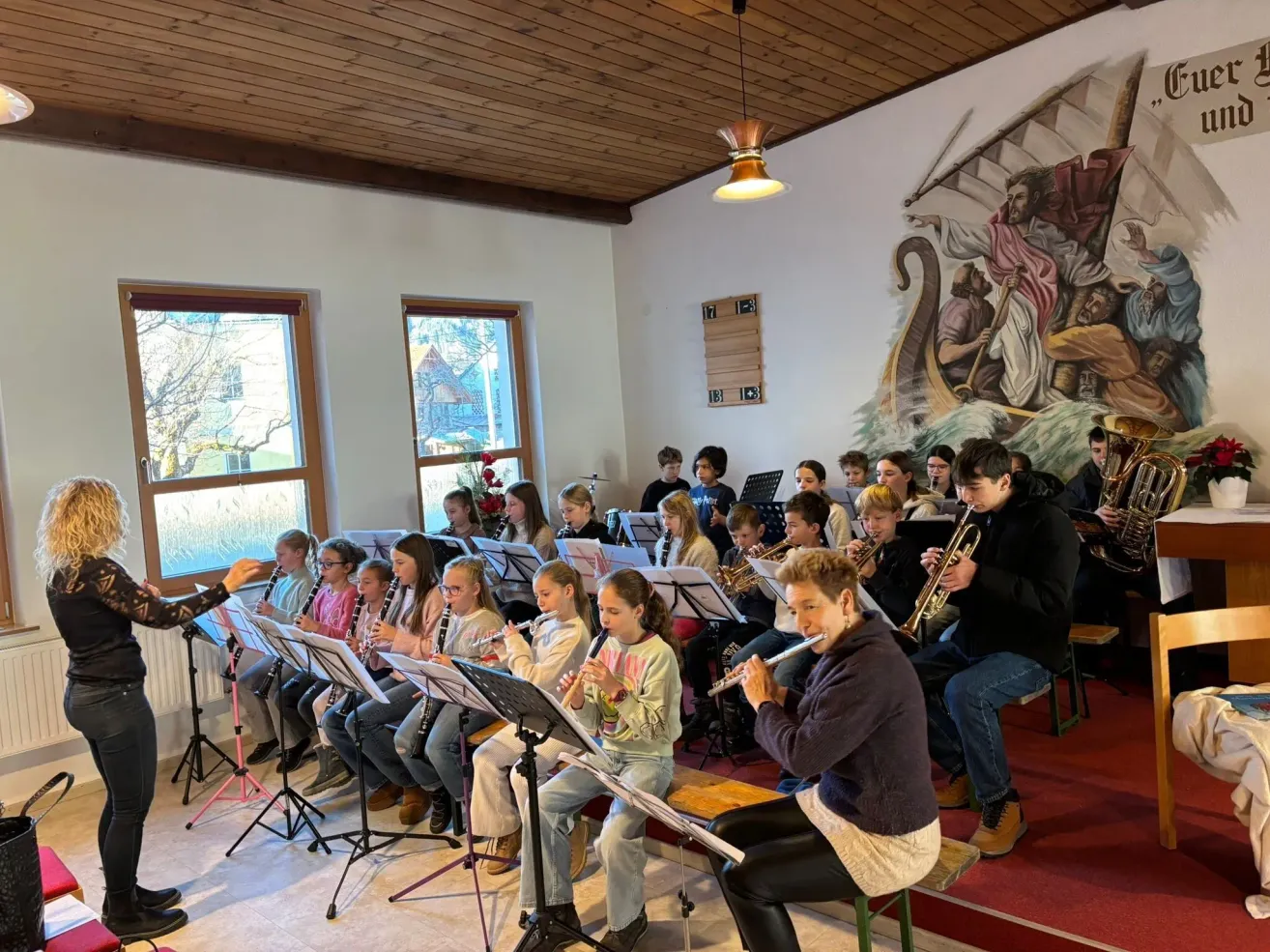 In einem Raum sitzen eine Gruppe von Kindern auf Stühlen und spielen Musikinstrumente, begleitet von einer Frau in einer braunen Jacke. An der Wand dahinter befindet sich ein großes Wandbild.
