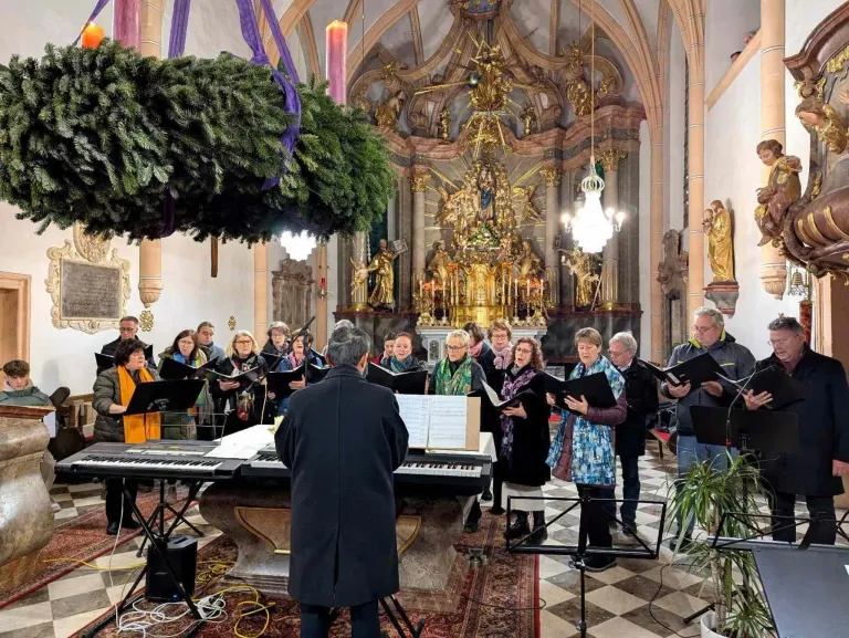 Ein Chor singt in einer Kirche mit Altar, goldenen Statuen und einer Kronleuchter. Die Chormitglieder singen und halten ihre Notenblätter. Der Dirigent steht vor einem Klavier. Die Kirche hat einen gemusterten Boden.