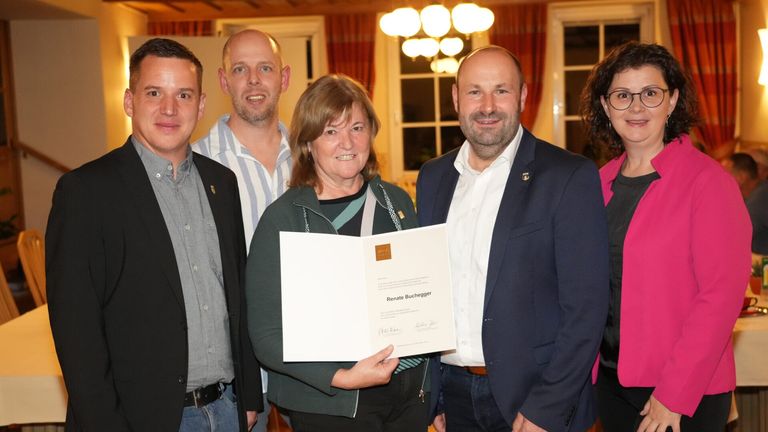 Eine Frau steht lächelnd vor einer Gruppe von vier Personen und hält eine Urkunde. Sie trägt eine grüne Jacke. Hinter ihr stehen zwei Männer in Freizeitkleidung, während ein weiterer Mann in einem Anzug die Urkunde hält. Sie befinden sich in einem Raum mit einer Kronleuchter und großen Fenstern.