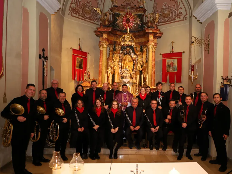 Eine Gruppe von Musikern in Schwarz und Rot steht vor einem kirchlichen Altar mit aufwendigen Verzierungen und religiösen Symbolen. Die Musiker halten verschiedene Blech- und Holzblasinstrumente.