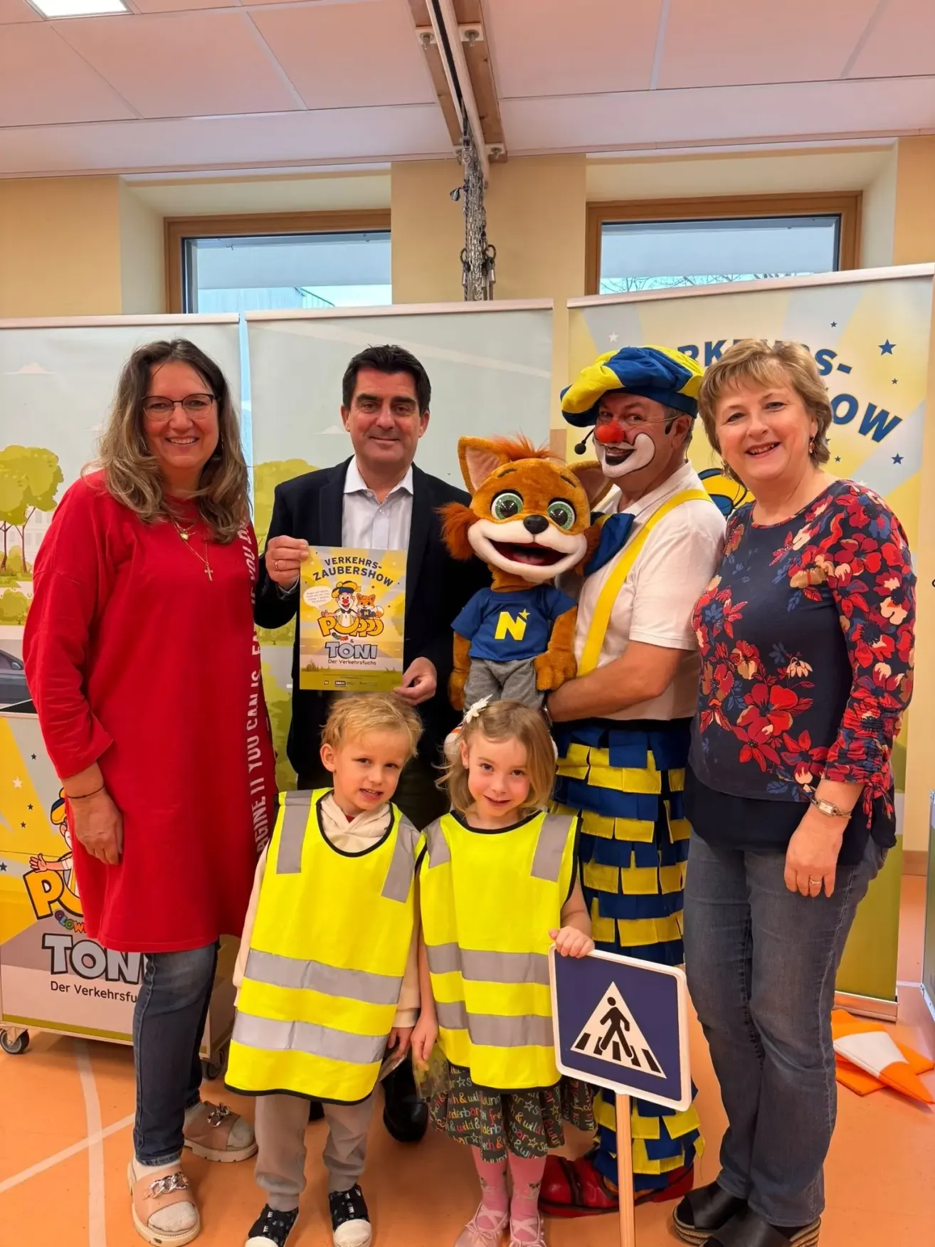 Eine Gruppe von Menschen posiert für ein Foto, darunter zwei Kinder in Sicherheitswesten, zwei Erwachsene und ein Clown in einer Fuchskostüm. Sie alle lächeln und stehen vor einem Hintergrund mit Text und einem Bild eines Baumes.