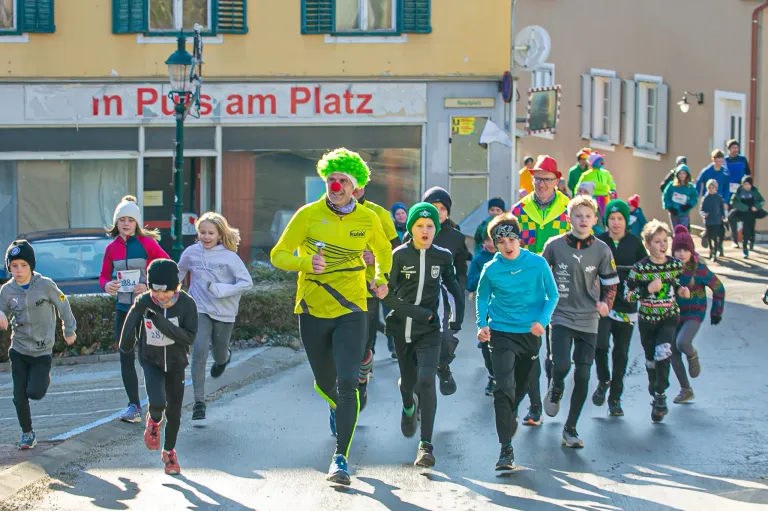Eine Gruppe von Menschen läuft einen Wettkampf, einer trägt eine Clownsnase. Sie laufen auf der Straße, und ein Mann vorne trägt ein gelbes Shirt. Hinter ihnen steht ein Gebäude mit einem Schild, das 'Pizzeria am Platz' besagt.