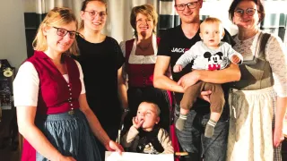 Eine Familie von sechs Personen in traditioneller Kleidung versammelt sich in einem Raum. Das Baby wird von einem Mann in einem schwarzen T-Shirt gehalten. Die Frau links hält ein Papier mit dem Wort 'Spende'.