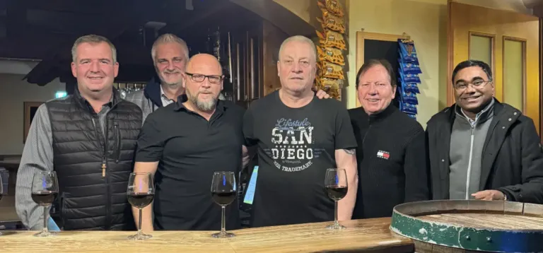 Vier Männer stehen hinter einer Bar und lächeln für ein Foto. Zwei sind in schwarzen Shirts, einer in der Mitte mit einem San Diego T-Shirt. Weingläser stehen auf der Bar.