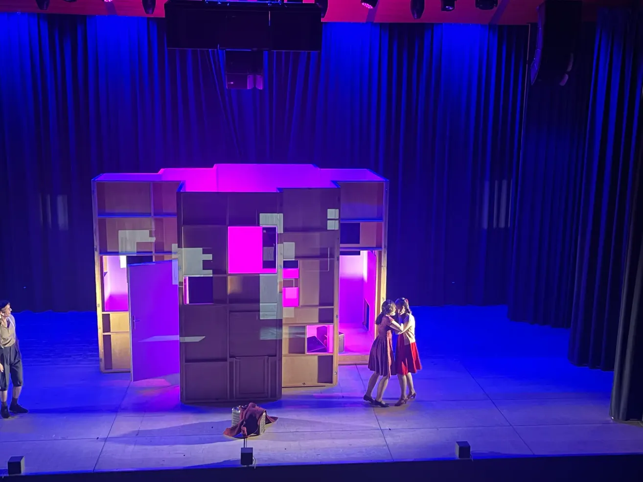 Zwei Frauen in einem roten und weißen Kleid stehen auf der Bühne und umarmen sich. Hinter ihnen steht ein großes Bühnenbild mit geometrischem Design, das von lila Lichtern beleuchtet wird.