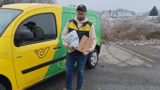 Ein Mann steht neben einem gelben und grünen Lieferwagen und hält eine Pappschachtel und einen Plastikbeutel in der Hand. Hinter ihm ist eine Person im Seitenspiegel des Lieferwagens zu sehen. Der Hintergrund zeigt eine Landschaft mit Bäumen und einem Haus.