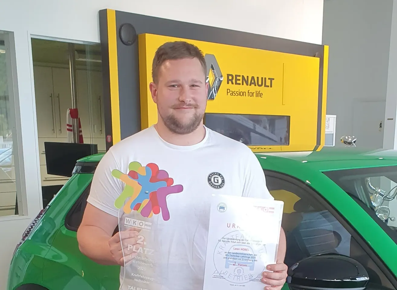 Ein Mann steht vor einem grünen Renault-Auto und hält einen Pokal und ein Zertifikat. Er trägt ein weißes T-Shirt mit einem farbenfrohen Design und einem Renault-Logo. Hinter ihm befindet sich ein gelbes Banner mit Renaults Slogan 'Passion für das Leben'.