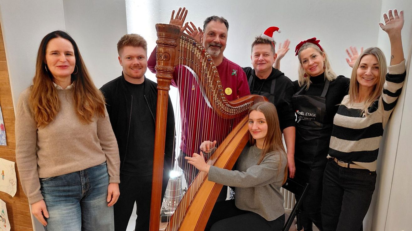 Eine Gruppe von Menschen posiert für ein Foto mit einer Frau, die eine Harfe spielt, eine Weihnachtsmütze trägt. Eine andere Frau winkt enthusiastisch. Der Mann im roten Shirt lächelt, während er den Hals der Harfe hält. Der Hintergrund ist eine einfache weiße Wand.
