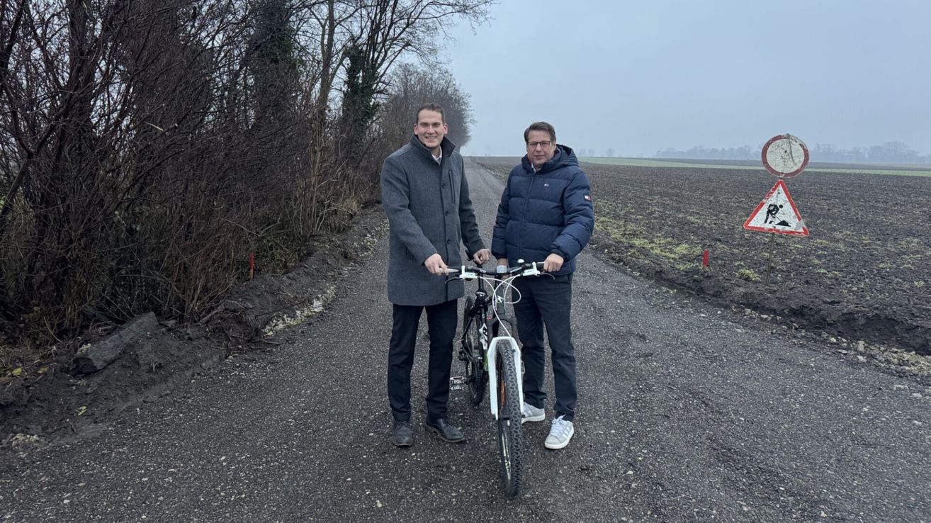 Zwei Männer stehen lächelnd auf einer Kiesstraße und halten ein Fahrrad zwischen sich. Einer trägt einen grauen Mantel, der andere einen blauen Jacke mit Logo. Bäume und Büsche säumen die Straße, im Hintergrund befindet sich ein Feld.