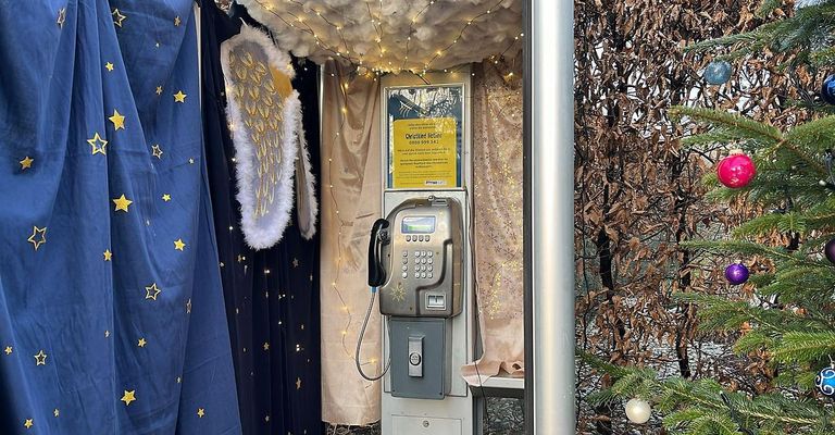 Eine Telefonzelle, dekoriert mit Engelsflügeln und Lichterketten, geschmückt mit einem festlichen Schild und Weihnachtsdekoration.