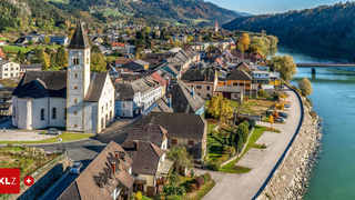 Ein Dorf mit einer Kirche, umgeben von Häusern, Grünflächen und Bergen, mit einem Fluss in der Nähe.