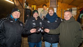 Vier Männer in Jacken halten Tassen mit einem schwarzen Design. Sie lächeln, und zwei haben Mützen. Hinter ihnen hält ein weiterer Mann eine Tasse. Es gibt Flaschen auf einem Regal, und ein Lautsprecher ist links.