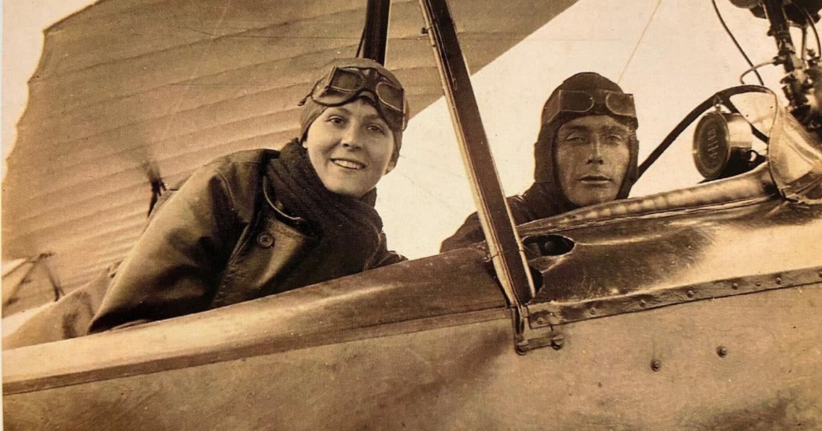 Zwei Piloten in einem frühen Flugzeug, einer lächelt, der andere sieht ernst aus. Beide tragen Schutzbrillen und Fluganzüge. Die Struktur des Flugzeugs ist sichtbar, mit einem offenen Cockpit.